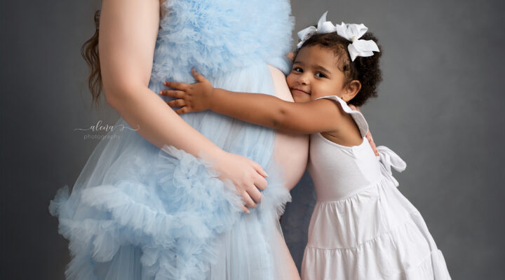 Young child hugging their pregnant mother’s belly during a maternity photoshoot, capturing the special bond between siblings before baby’s arrival